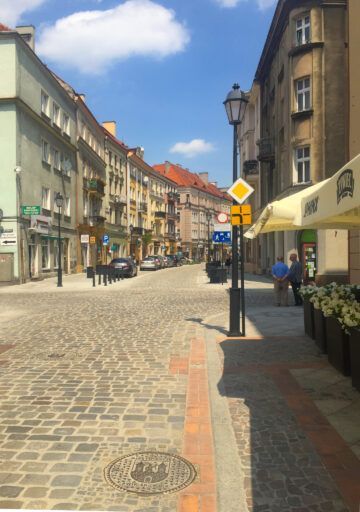 Ulica w centrum Kalisza z oprawą oświetleniową 01D Vera zamontowaną na stylizowanym słupie latarni. Oprawa wkomponowana w historyczną zabudowę, stanowi element miejskiego oświetlenia w strefie pieszej.