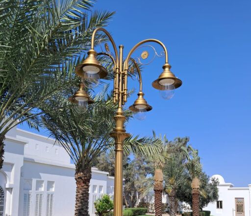 Bogato zdobiona latarnia uliczna w złotym odcieniu z czterema oprawami 04/Aries/A, na tle palm i białych budynków w słoneczny dzień